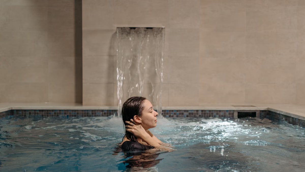 Adult woman relaxing in an indoor swimming pool near a cascading waterfall.